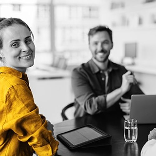 Female,Designer,Smiling,At,The,Camera,While,Sitting,At,A Female designer smiling at the camera while sitting at a table with her team. Business woman having a meeting with her colleagues in an office.